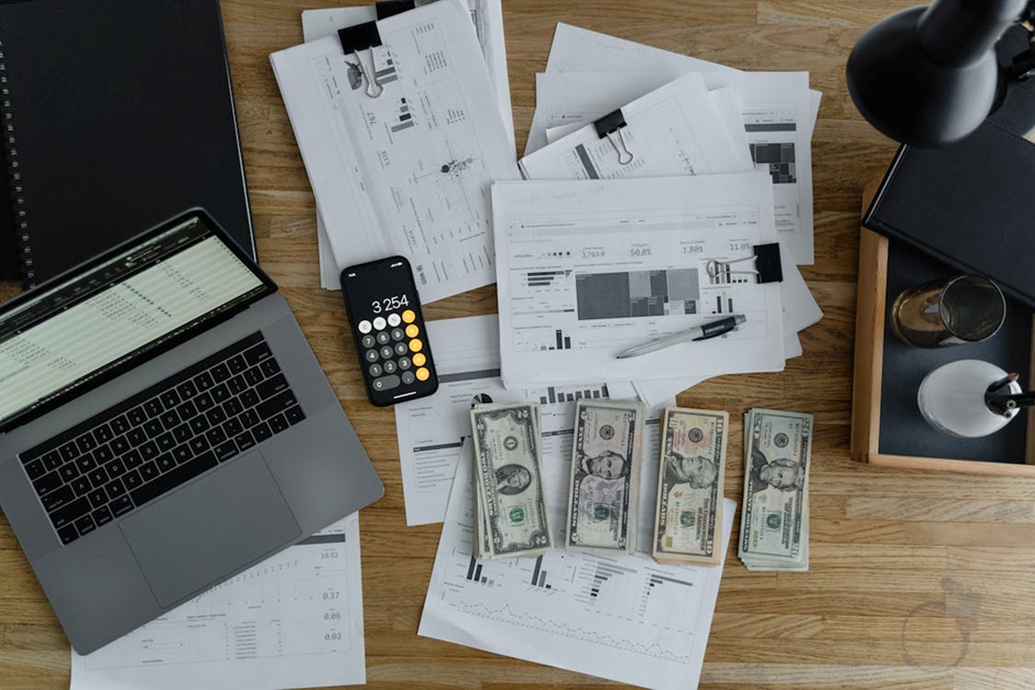 An image of a laptop, calculator, paperwork, and stacks of dollar bills on a table