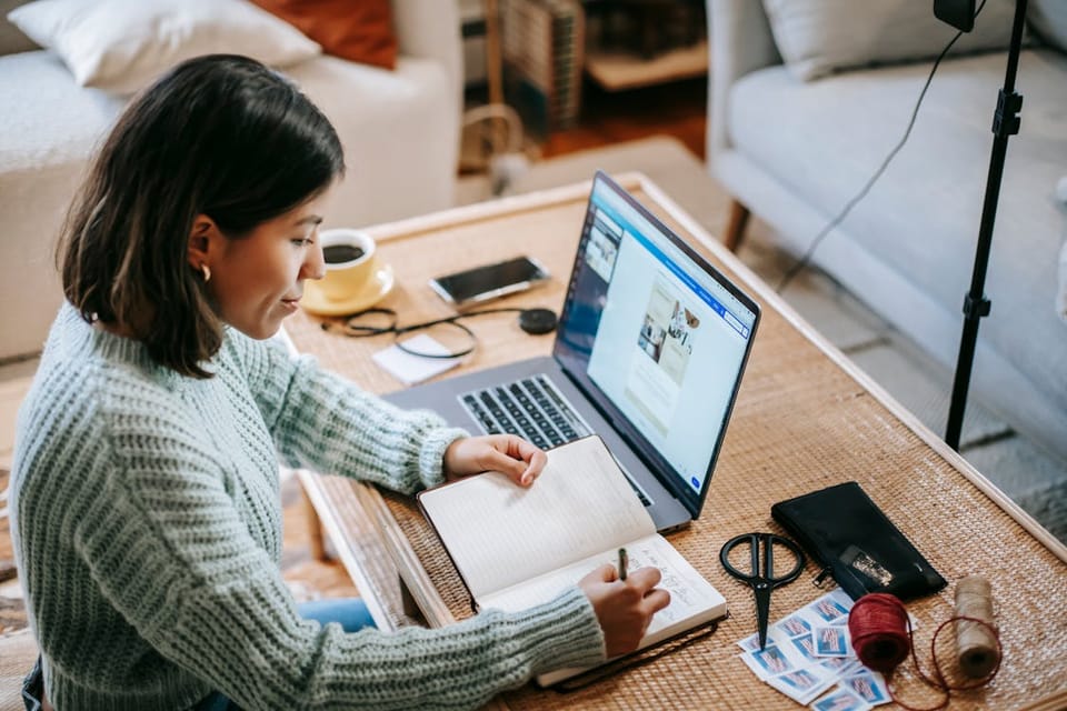 A woman sitting at a desk opposite a laptop and a filming rig. She is writing in a notebook